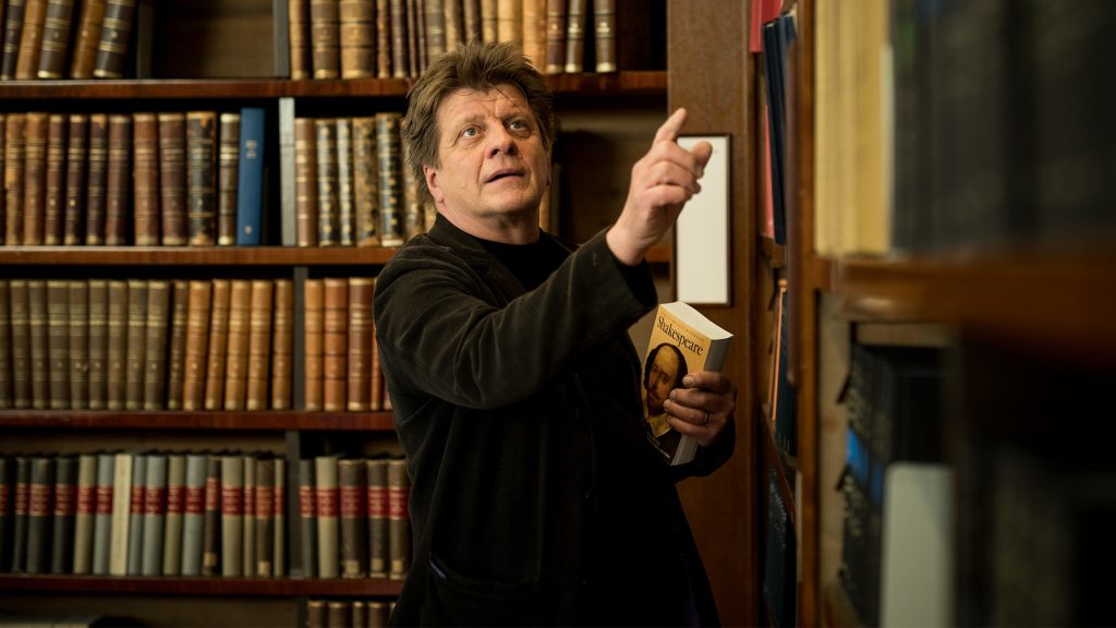 Henkilö osoittamassa jotain kirjaa kirjahyllyllä. Hänellä on kädessä Shakespearen teosten kokoelma.