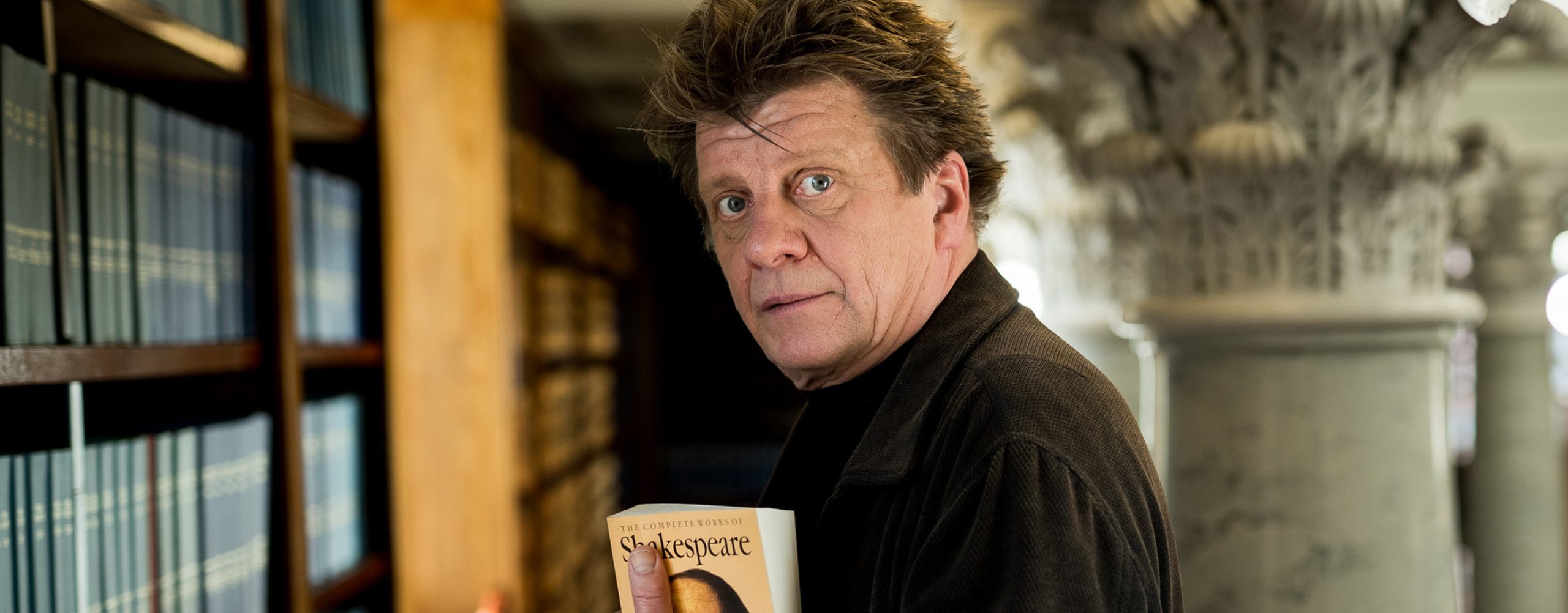 Henkilö katsomassa kameraan mustassa yläosassa. Hänellä on kädessä Shakespearen teosten kokoelma.