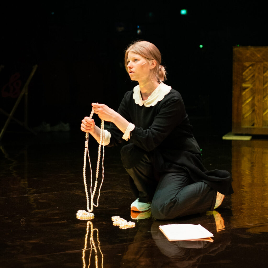 Näyttelijä Roosa Söderholm polvillaan teatterin näyttämöllä, kädessä pitkä helminauha.