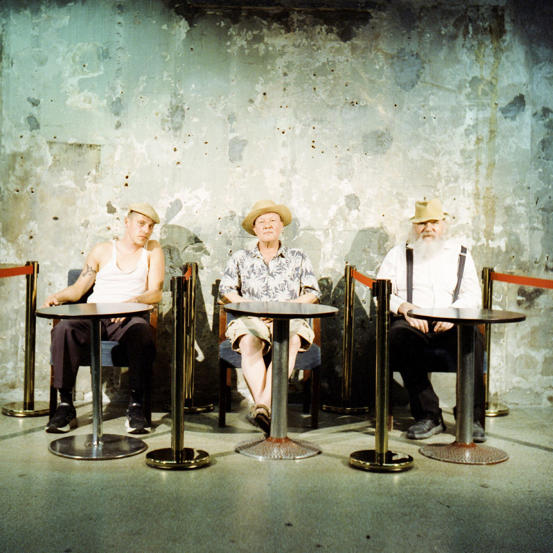 Three people sit by tables in front of a concrete wall and look into the camera.