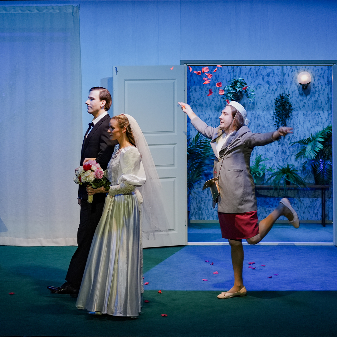 A man in a wedding suit walking beside a woman in a wedding dress holding a bouquet of flowers. Behind them, a person throws red petals for the couple while standing with one leg raised.