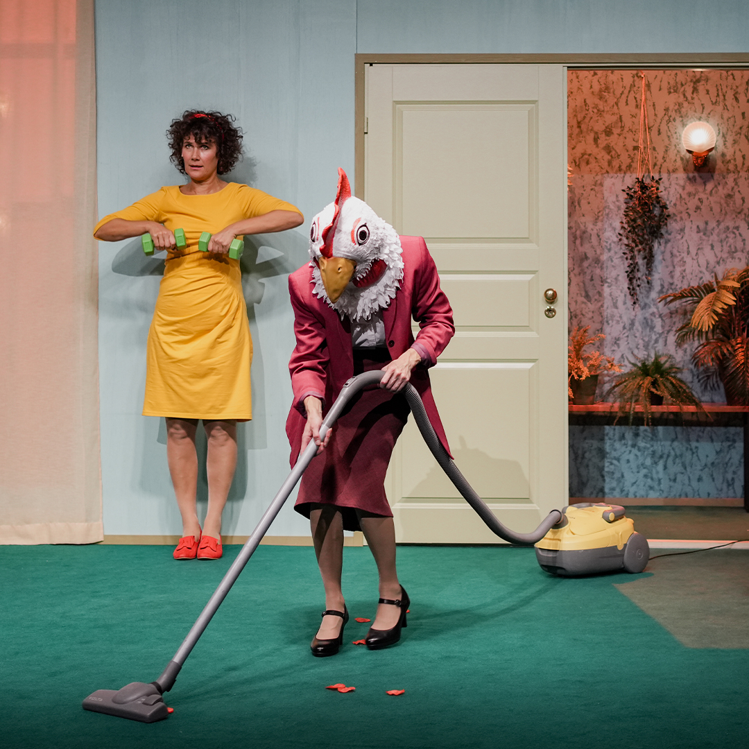 A woman in a yellow dress lifting a pair of dumbbells and another woman in a red blazer wearing a chicken mask vacuuming red petals from the floor.