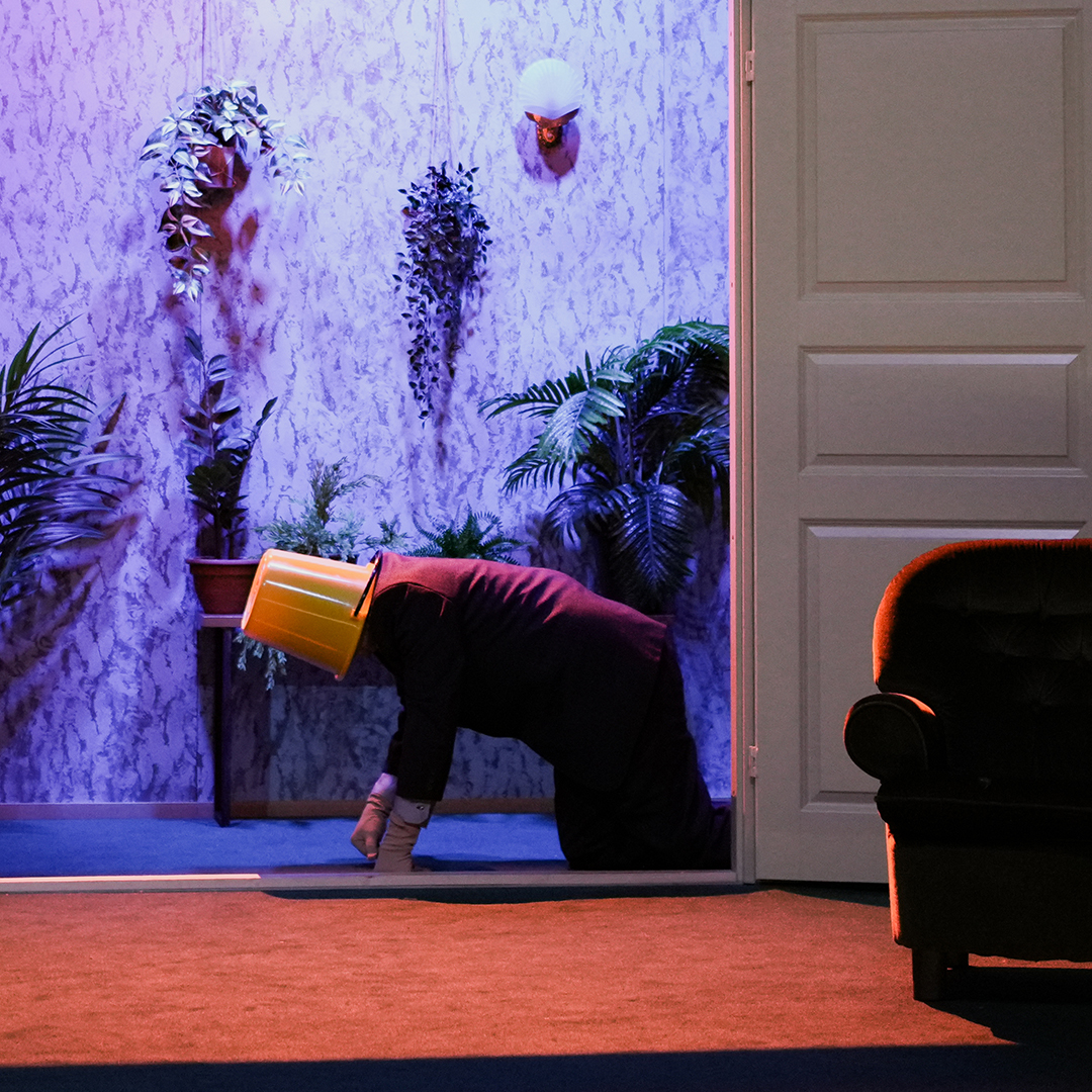 An actor crawling on the floor through a doorway wearing a red blazer and a yellow bucket on its head.
