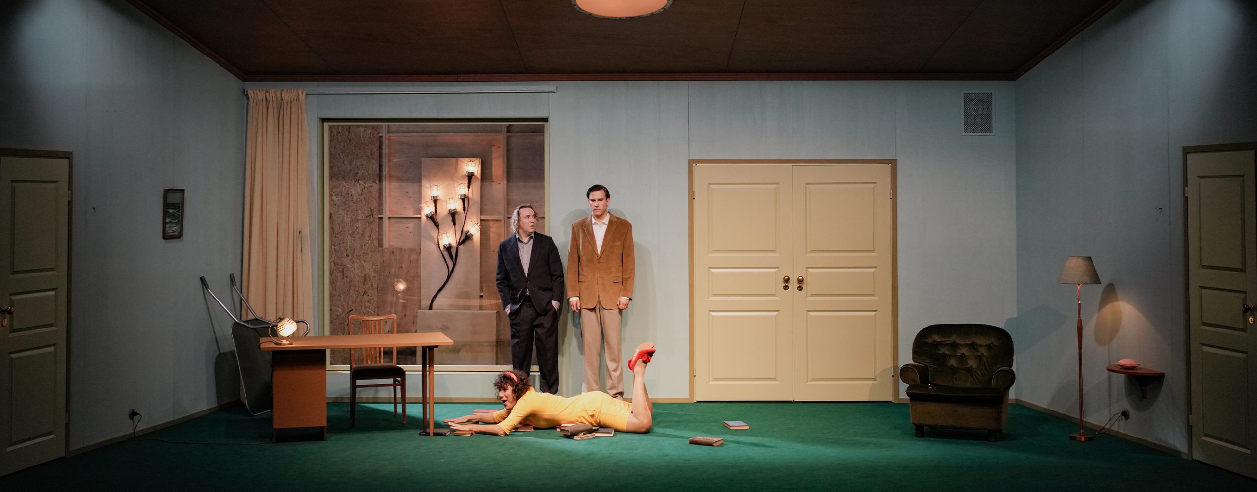 A wide angle shot of the stage with three actors in the middle. Two men are standing close to each other wearing black and brown suits and in front of them is a woman wearing a yellow dress and red shoes laying face down on the floor with books.