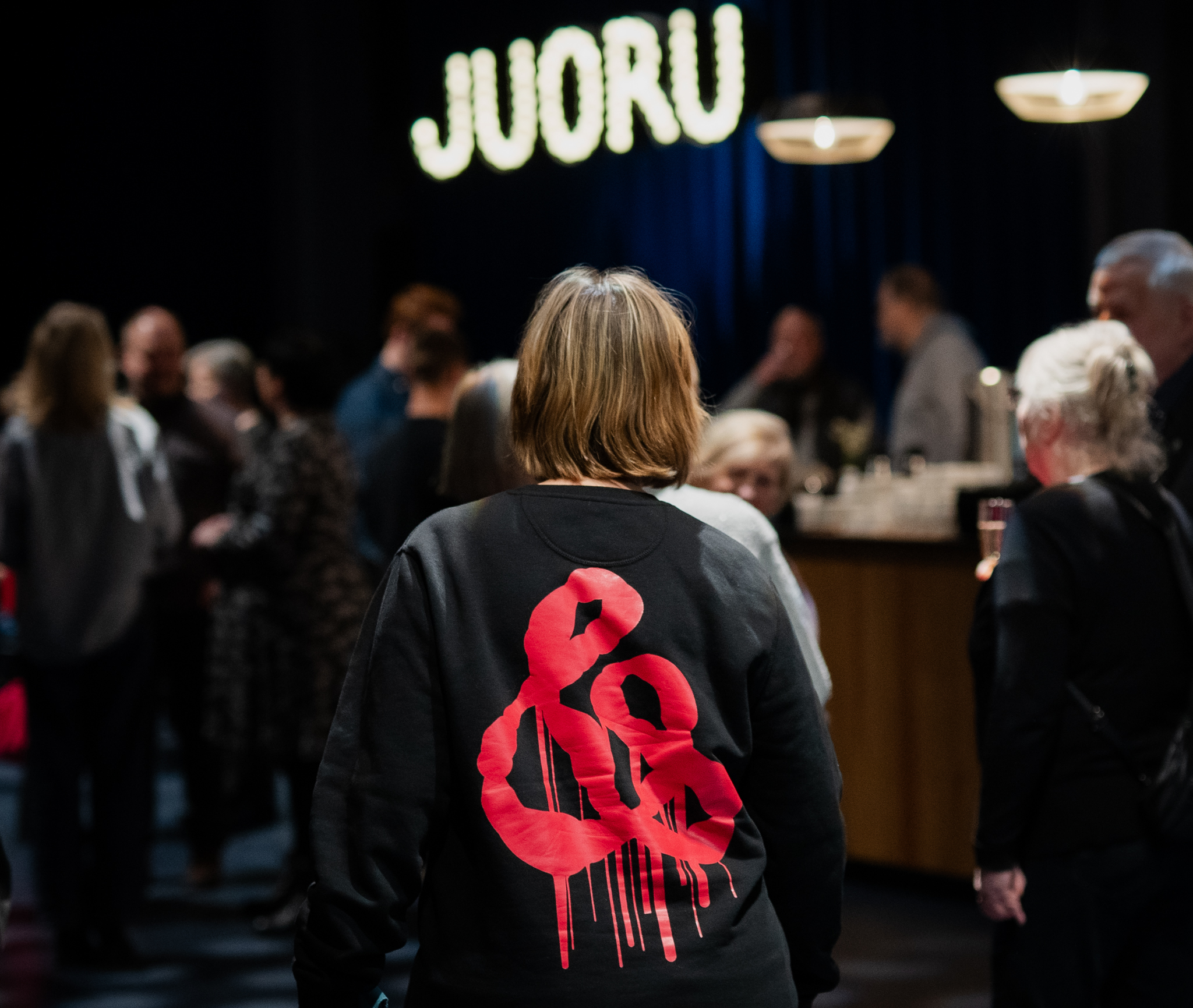 Teatterin lämpiössä ihmisiä, taustalla Bar Juorun valomainos, keskellä henkilö, jonka paidan selässä suuri punainen &-logo.