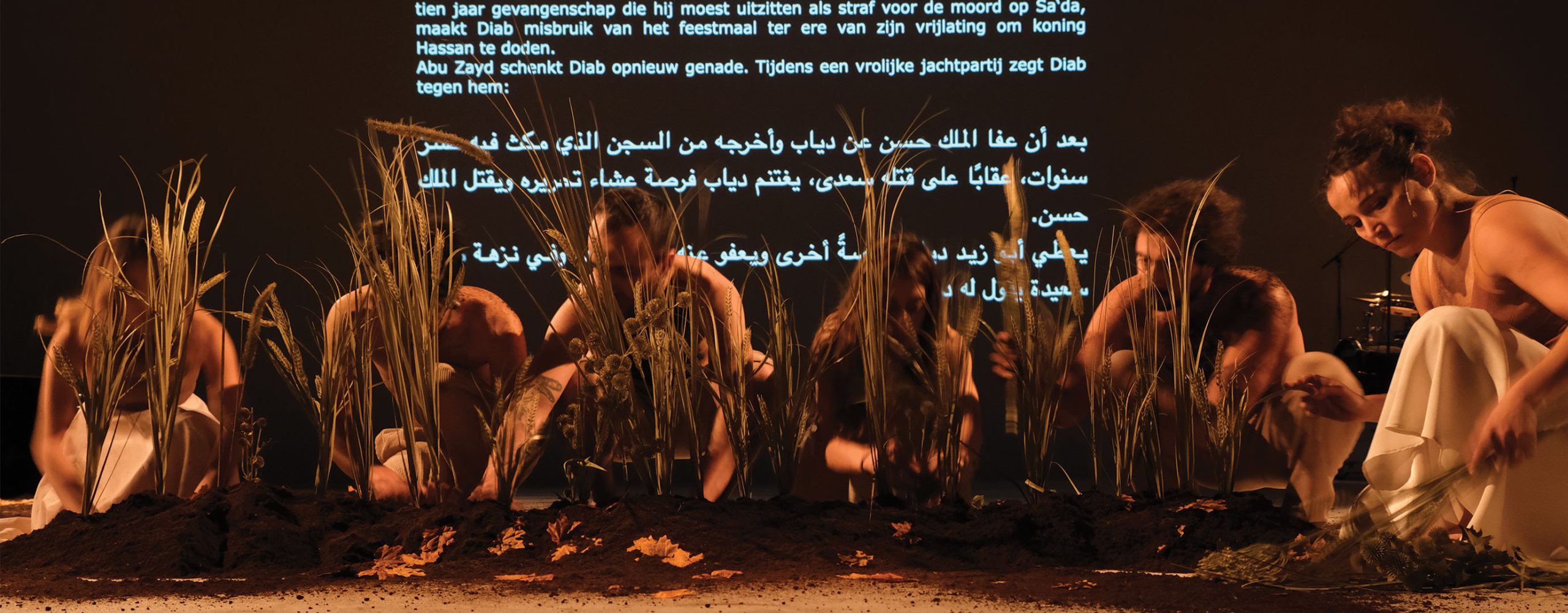 Kuusi henkilöä teatterin näyttämöllä kyykyssä ruohikon takana. Taustalla tekstiä hollanniksi ja arabiaksi.